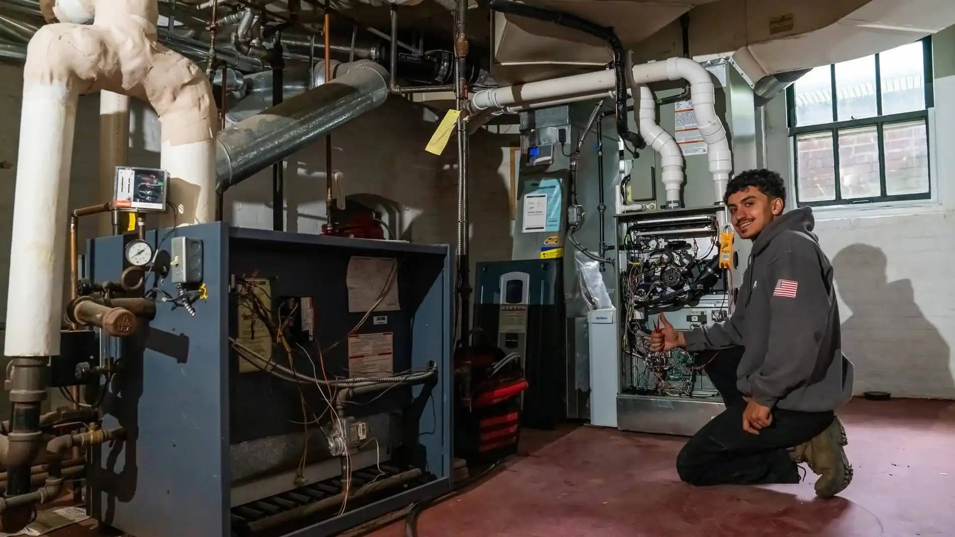 Raber technician kneeling at an opened high efficiency furnace in a basement, with an older cast iron boiler still piped in along the far wall, photographed on a Raber job in Northeast Ohio
