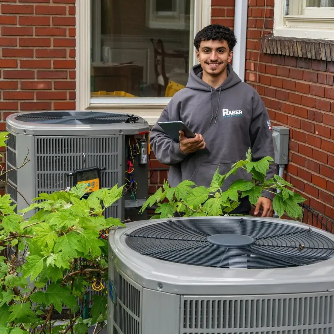 Raber technician holding a tablet between two outdoor air conditioning condensers at the side of a brick house