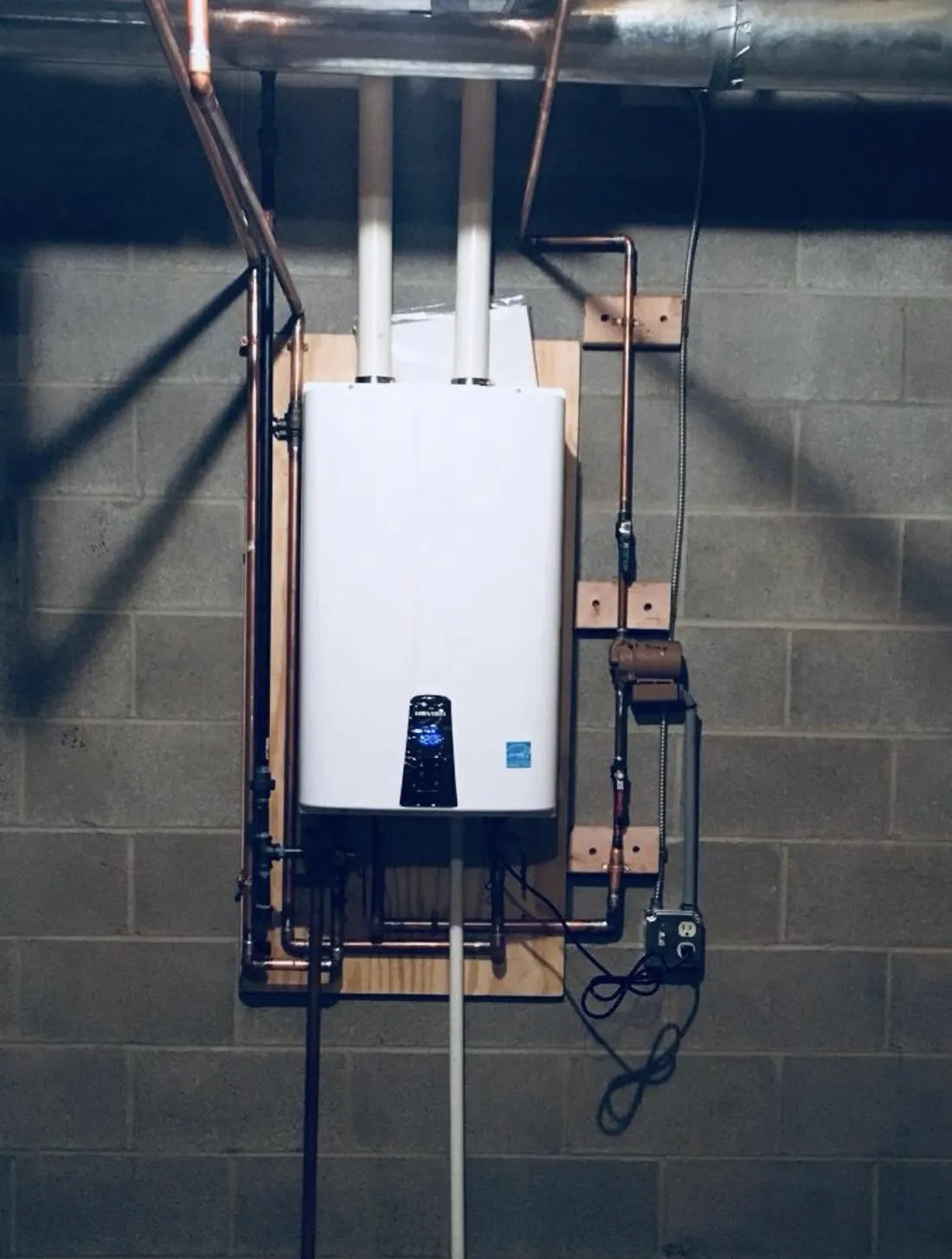 Tankless water heater mounted on a plywood backer against a block basement wall, with twin vents and copper supply lines run to it