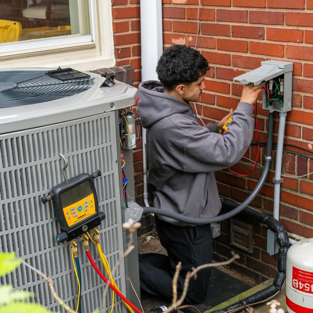 Raber technician checking the wiring in an outdoor disconnect while a refrigerant manifold reads pressures on the condenser beside him, photographed on a Raber job in Northeast Ohio