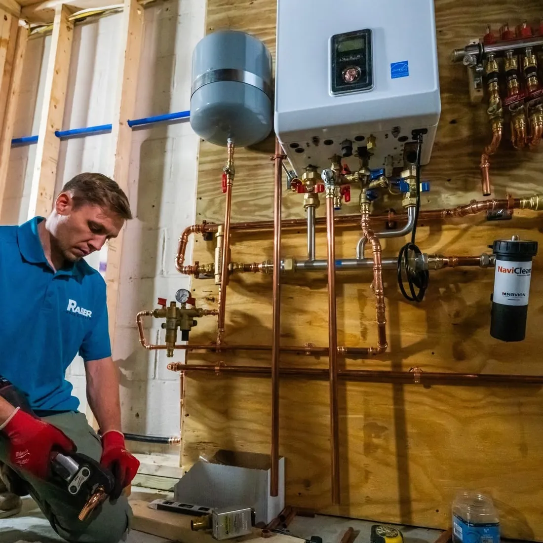 Raber technician kneeling at a newly installed combi boiler with the copper manifold, isolation valves and system filter piped in above, photographed on a Raber job in Northeast Ohio