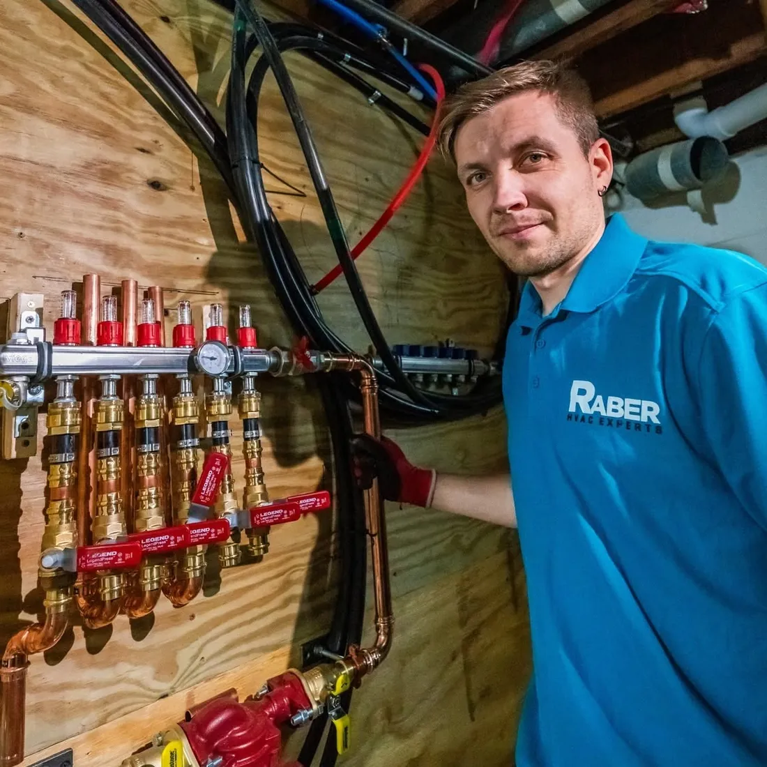Raber technician beside a stainless hydronic manifold with six flow meters, brass isolation valves and press fit copper supply lines on a plywood backer, photographed on a Raber job in Northeast Ohio